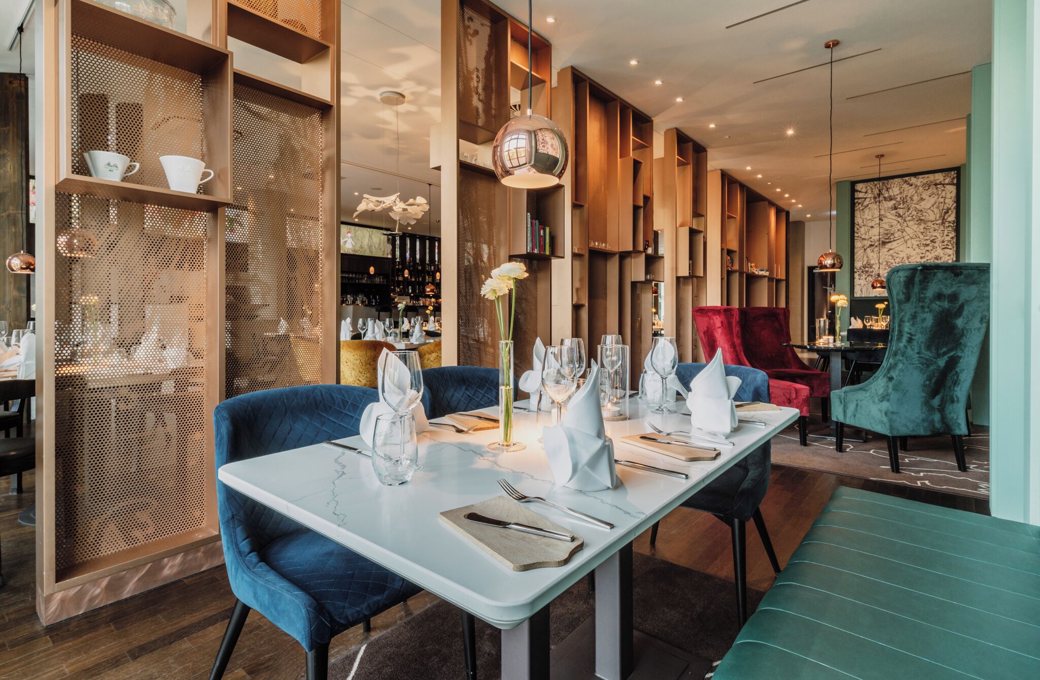 Een restaurant met een modern interieur, met een lange eettafel, blauwe gestoffeerde stoelen en een bar op de achtergrond.