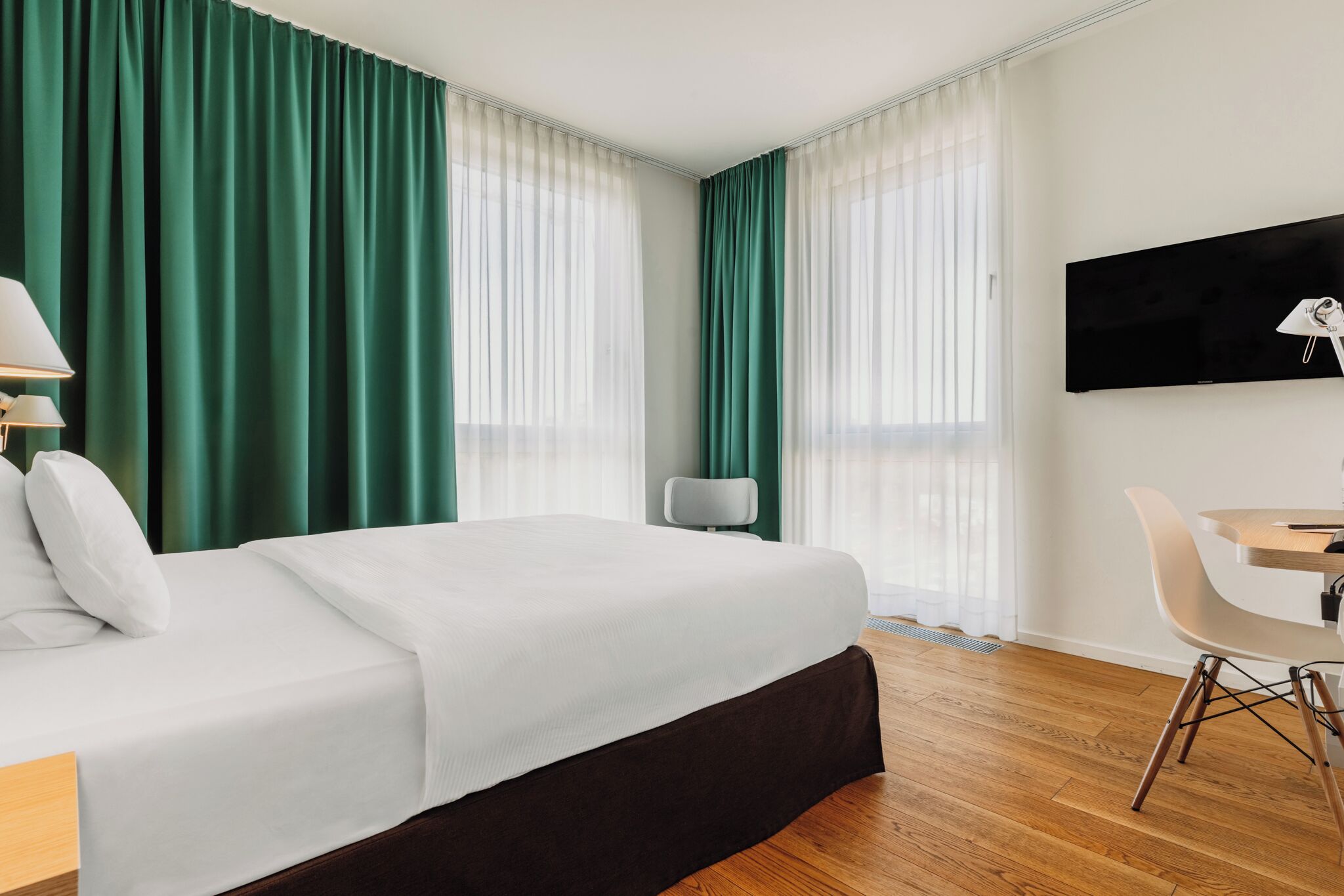 A hotel room with a bed, a desk, a chair, a TV, and green curtains.