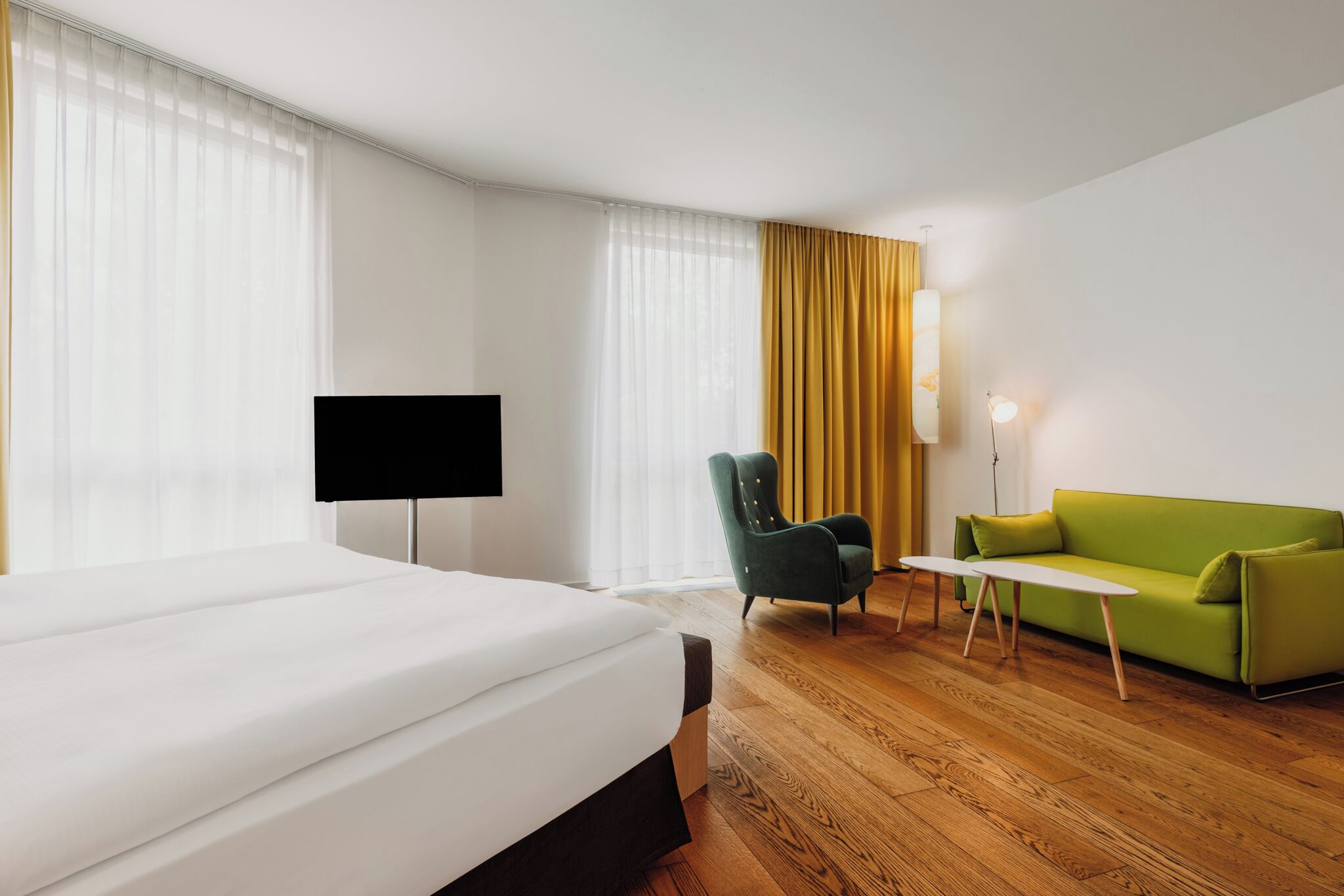 Quarto de hotel com cama, sofá verde, cadeira, televisão e abajur.