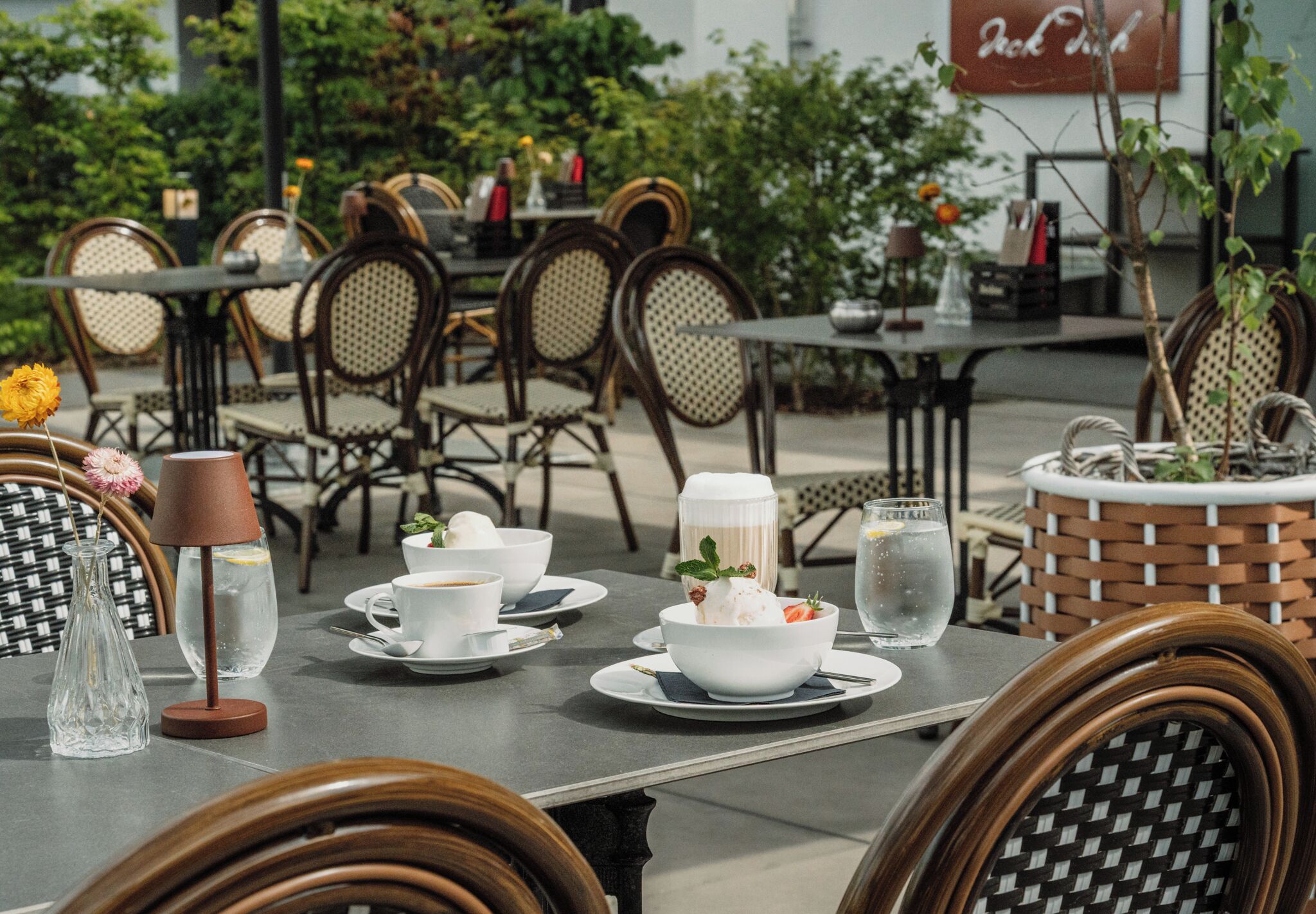 Un área de comedor al aire libre con sillas de mimbre, mesas dispuestas con platos blancos, tazas y un bol de postre.