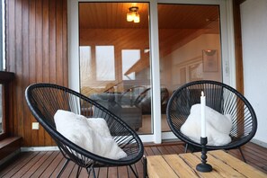 Deux fauteuils en osier noir avec des coussins blancs, placés sur une terrasse en bois, une porte-fenêtre coulissante donnant sur un espace intérieur et un plafond en bois avec un luminaire.