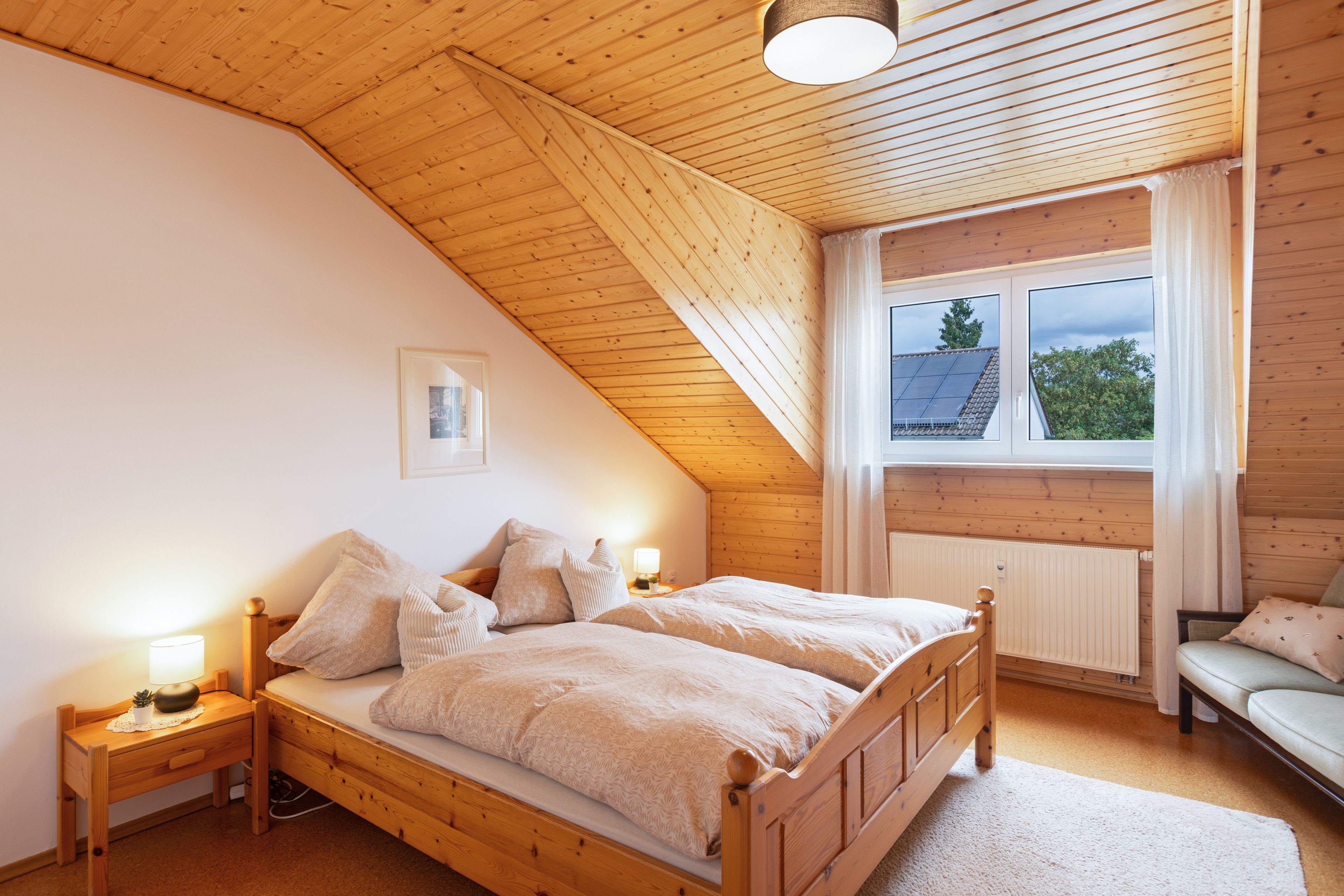 Ein Schlafzimmer mit geneigter Decke, einem Bett mit Holzrahmen, einem Nachttisch mit Lampe, einem Fenster mit Vorhängen und einer Heizung.