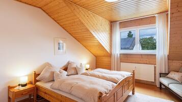 Ein Schlafzimmer mit geneigter Decke, einem Bett mit Holzrahmen, einem Nachttisch mit Lampe, einem Fenster mit Vorhängen und einer Heizung.