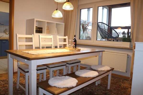 Un coin repas avec une table en bois, des chaises blanches, un banc agrémenté de coussins moelleux et une vue sur l’extérieur par une fenêtre.