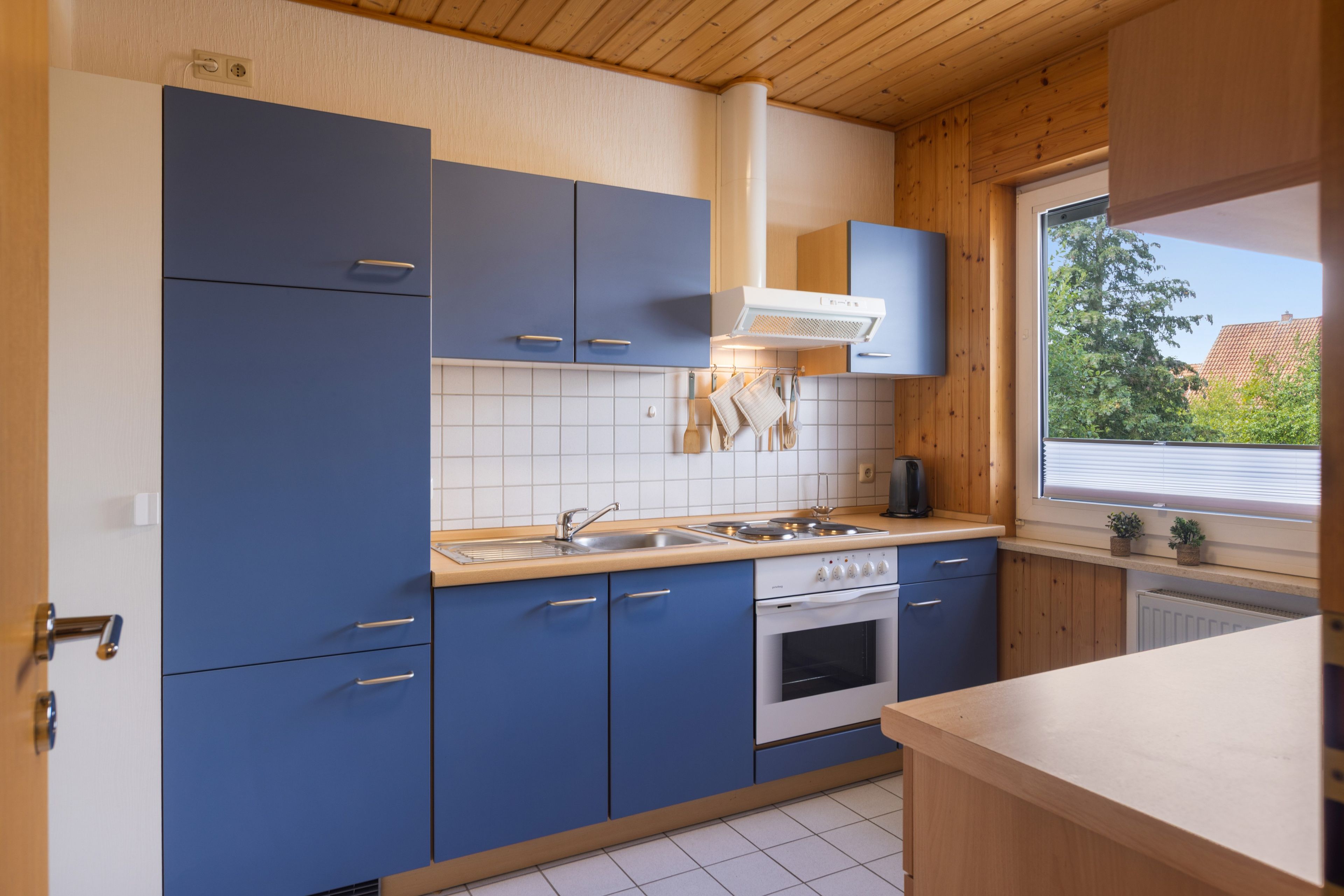 Een moderne keuken met blauwe kastjes, een witte oven en een raam met uitzicht op groen.