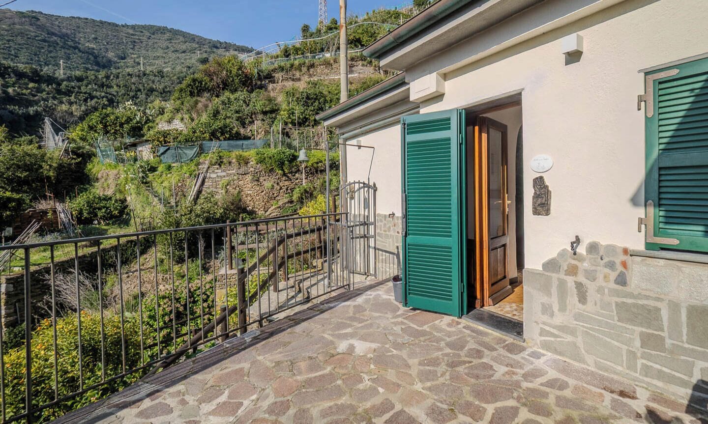 A stone-floored balcony with a metal railing, a house with green shutters, and a view of a hillside.