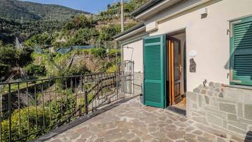 A stone-floored balcony with a metal railing, a house with green shutters, and a view of a hillside.