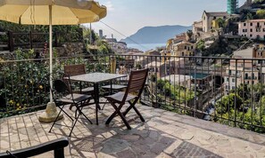 A terrace with a table and chairs, a yellow umbrella, and a view of a hillside town. - Camere Giuliano Basso (Vernazza)