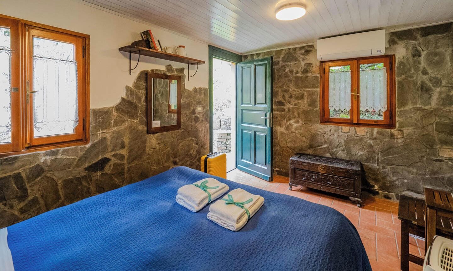 A room with a stone wall, a bed with blue bedding, a wooden dresser, and a blue door.