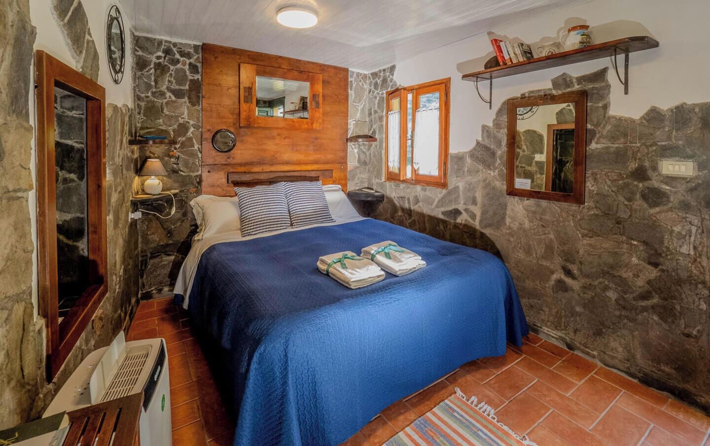 Une chambre avec un mur en pierre, un lit recouvert d’une couverture bleue, une tête de lit en bois, un miroir et une étagère remplie de livres.