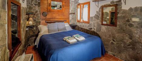 Une chambre avec un mur en pierre, un lit recouvert d’une couverture bleue, une tête de lit en bois, un miroir et une étagère remplie de livres.