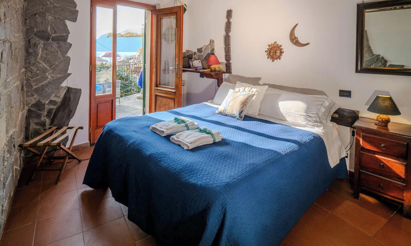 A bedroom with a bed, a wooden dresser, a nightstand, a lamp, and a view of the sea through a door.