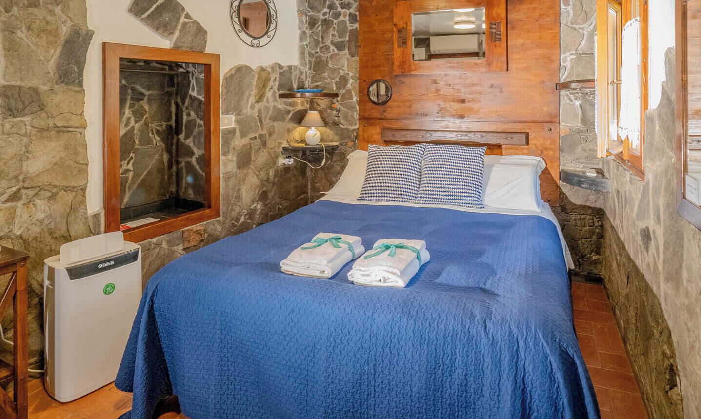 Una stanza con parete in pietra, un letto con copriletto blu, due asciugamani, un termosifone e una testiera in legno.