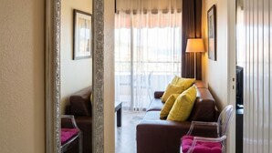 Een hotelkamer met een grote spiegel, een bruine bank met gele kussens, een transparante plastic stoel, een lamp en een televisie.