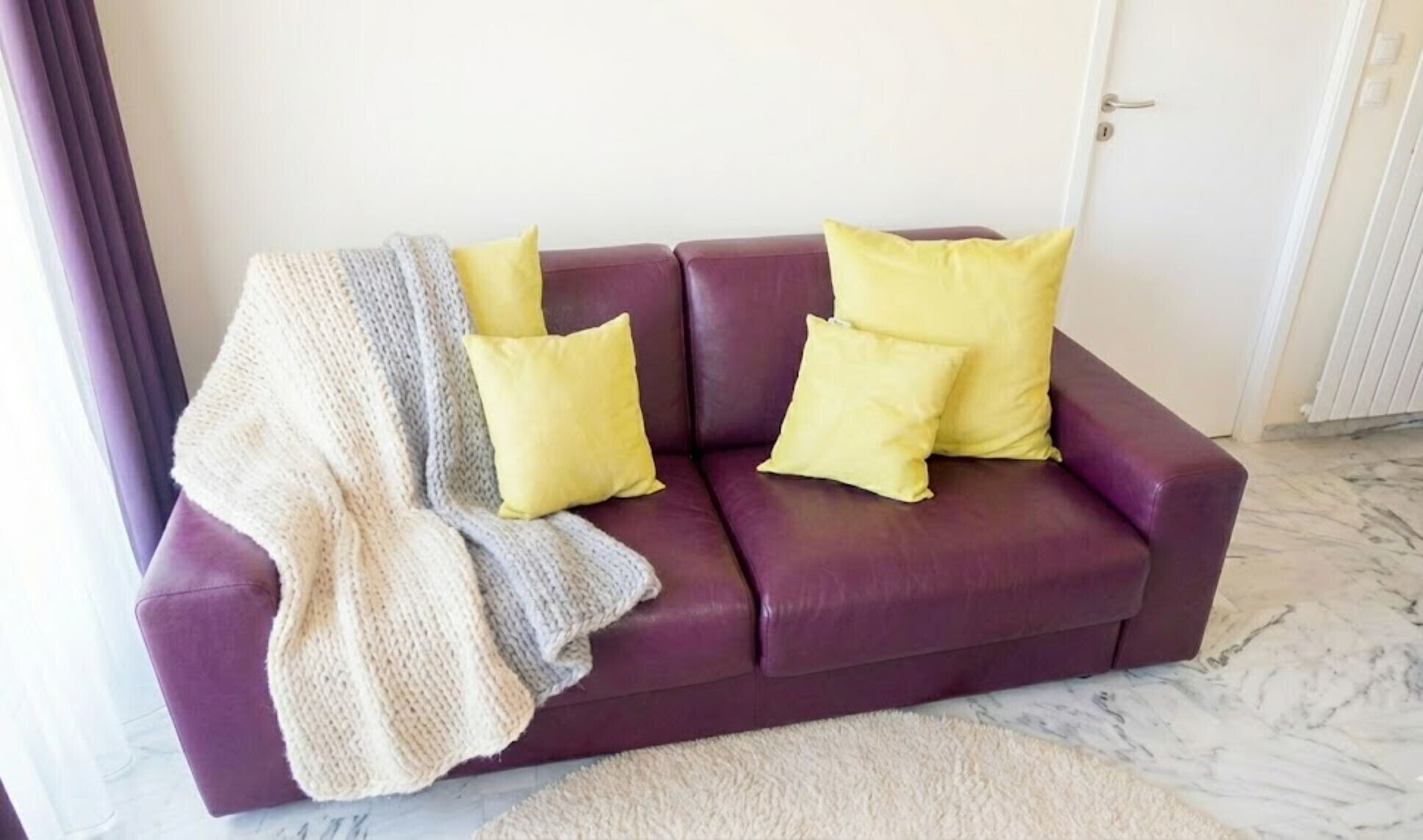 A purple sofa with three yellow cushions and a knitted throw blanket.