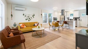 En moderne stue med sofa, sofabord og stole, et køkken med hvide skabe og et spiseområde, samt et stort vindue der giver naturligt lys.