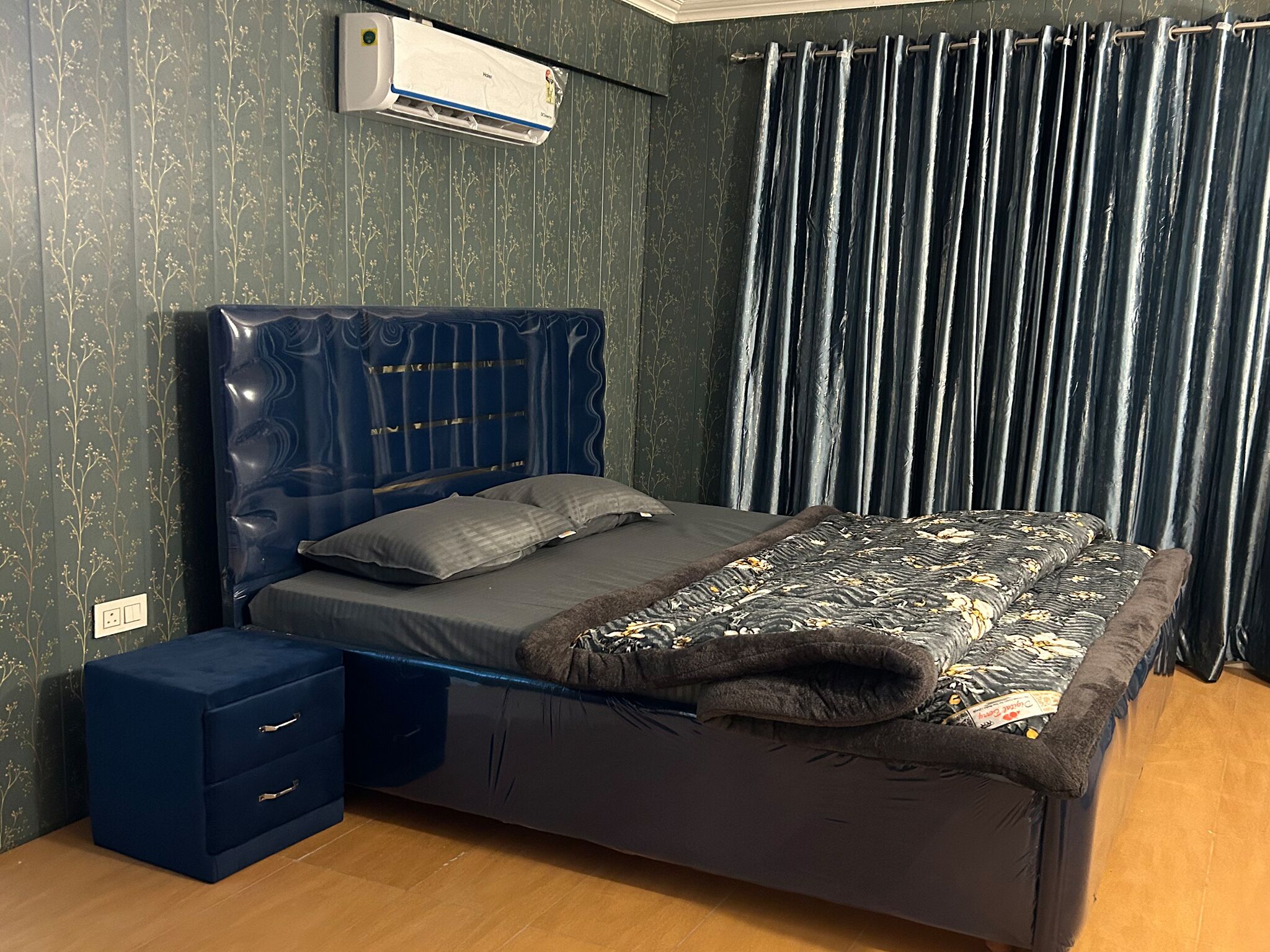Een slaapkamer met een bed, een nachtkastje, een dressoir, een wandairconditioner en een raam met gordijnen.