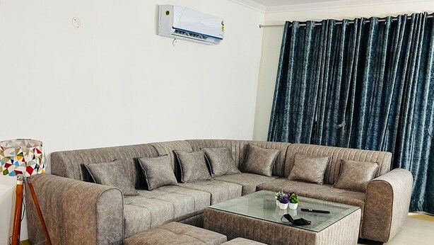 A living room with a sectional sofa, ottomans, and a glass coffee table. - Stay Royal Landran (Sahibzada Ajit Singh Nagar)