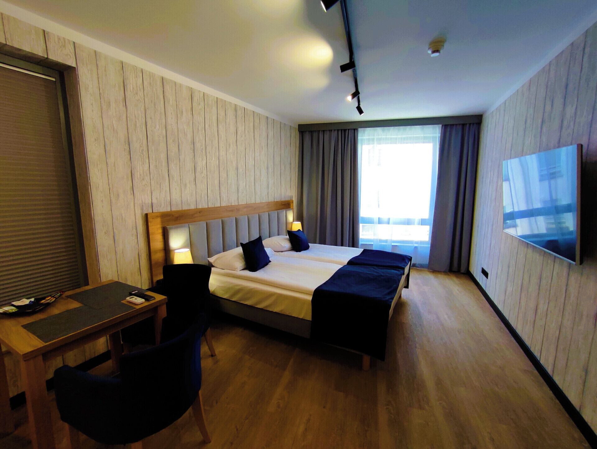 A hotel room with a bed, two bedside tables, a desk, a chair, a TV, and a window with curtains.