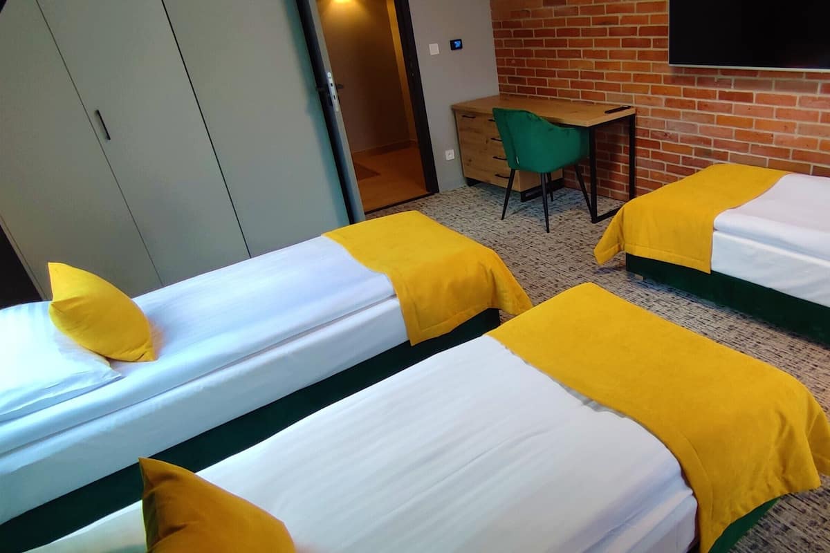 A room with two beds, each with a yellow blanket and pillow, a desk with a chair, and a brick wall.