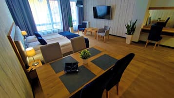 Una habitación de hotel moderna con una cama grande, un área de comedor con una mesa y sillas, un televisor montado en la pared y un balcón con cortinas.