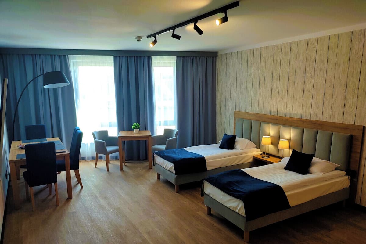 A hotel room with two beds, a desk with a chair, a small table with a plant, and a lamp.