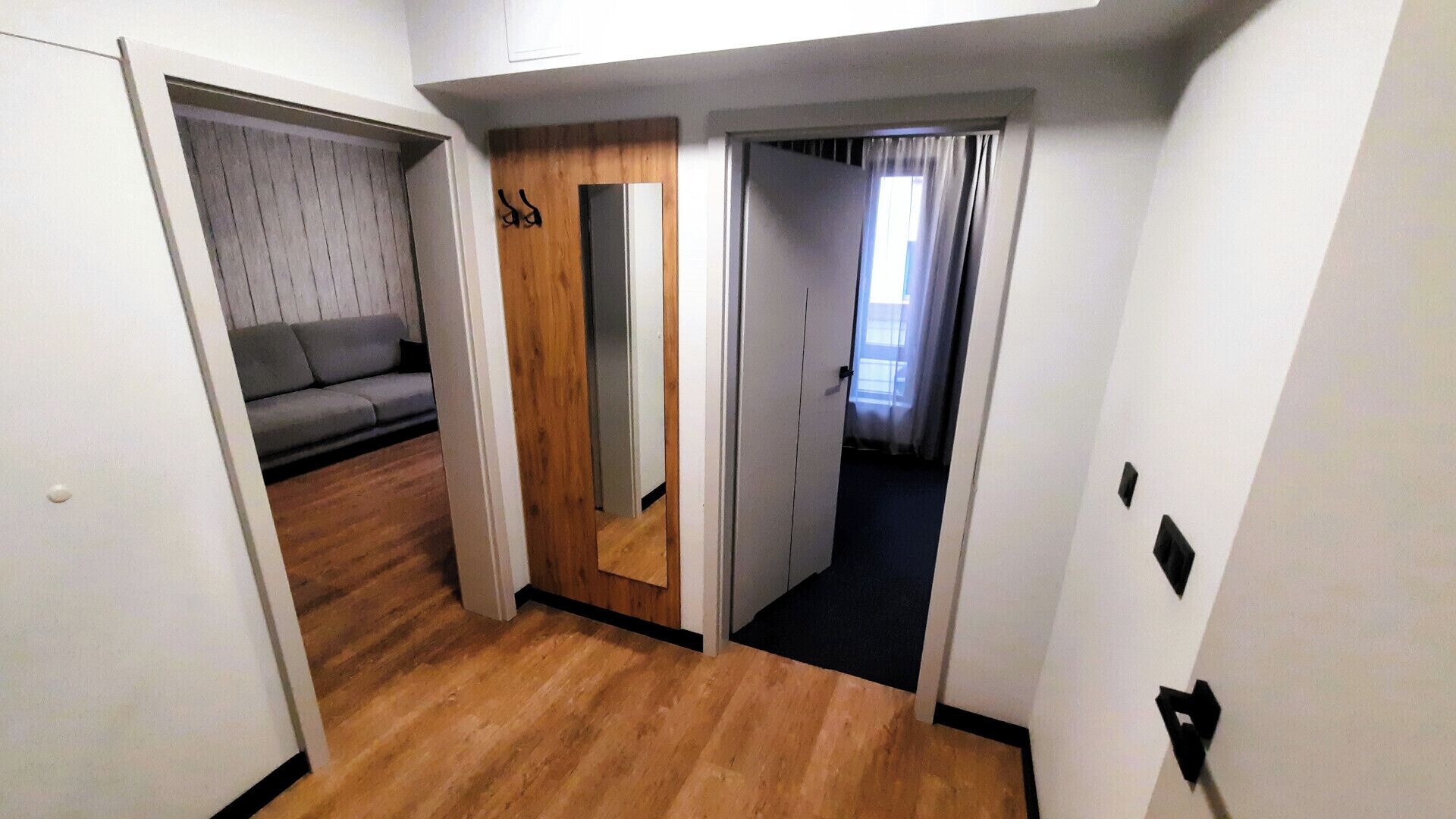 Ein schmaler Flur mit Holzdielen, ein graues Sofa im linken Raum und eine Tür mit Holzverkleidung.