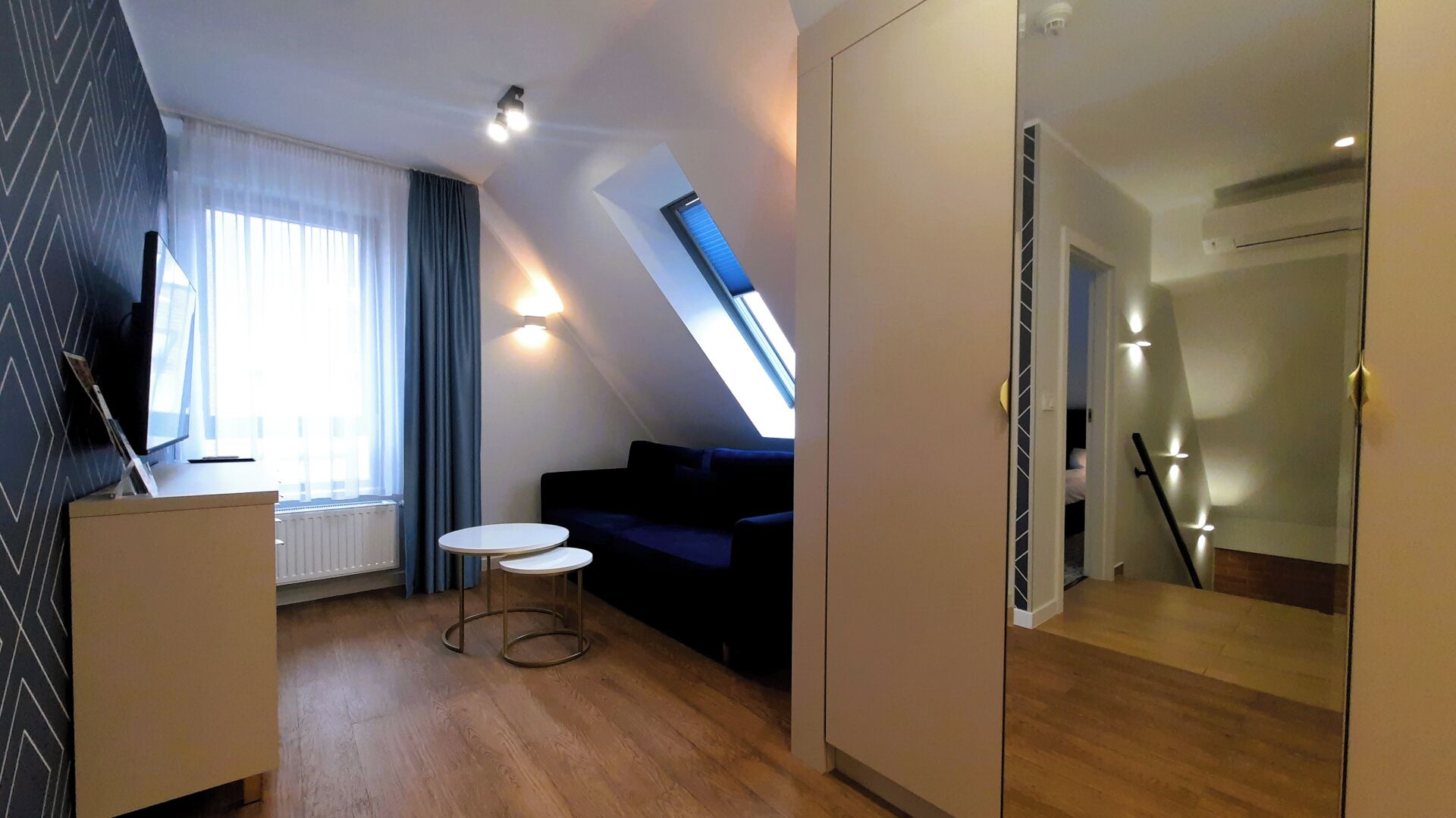 A modern, well-lit room with a sofa, a small table, and a TV on a stand. There is a window with curtains, a skylight, and a staircase leading to another level.