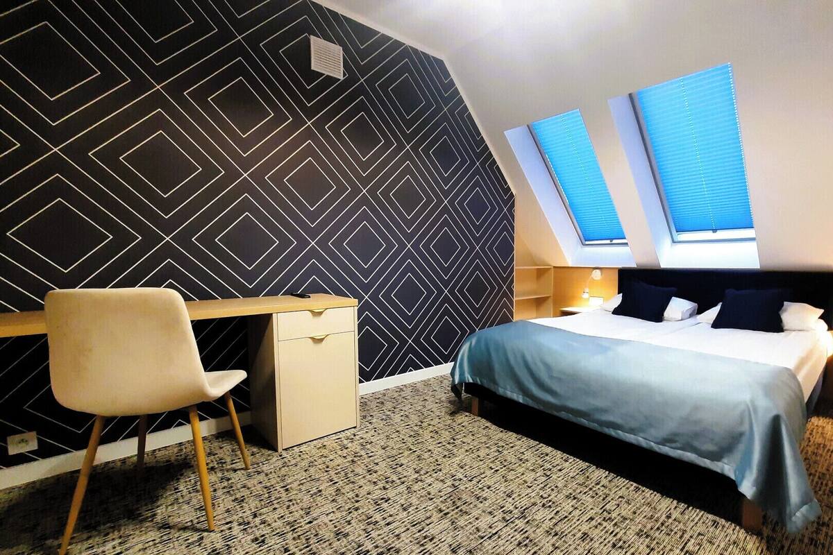 A hotel room with a geometric patterned wall, a desk with a chair, a bed with two pillows, and a skylight.