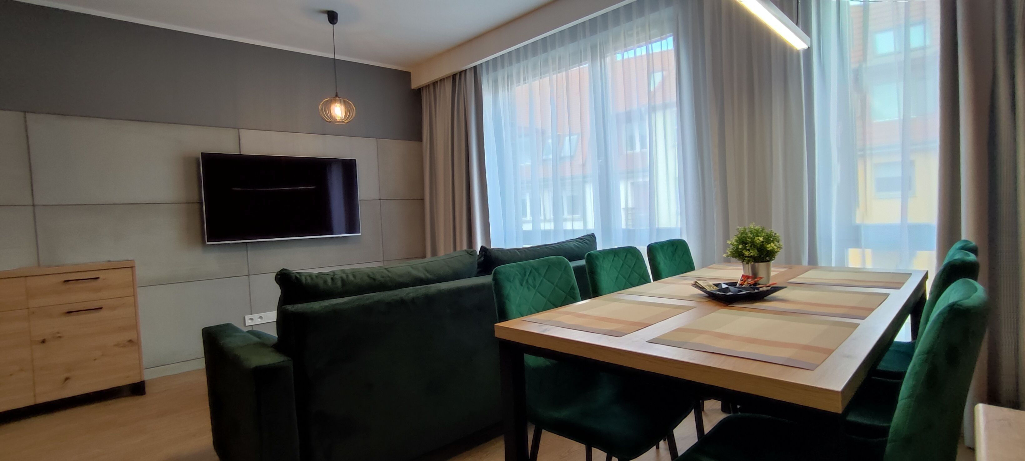 Una sala moderna con un televisor de pantalla plana montado en la pared, un sofá verde, una mesa de comedor con sillas y un amplio ventanal con cortinas translúcidas.