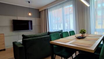Una sala moderna con un televisor de pantalla plana montado en la pared, un sofá verde, una mesa de comedor con sillas y un amplio ventanal con cortinas translúcidas.