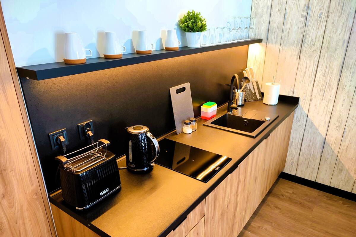 A modern kitchen with a black countertop, a coffee maker, a kettle, a sink, and a shelf with cups and a plant.