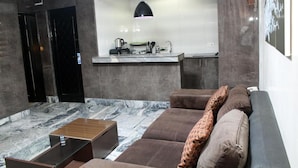 A modern living room with a marble floor, a sofa with patterned cushions, a coffee table, and a kitchen area with a black pendant light. - The Regal Boutique Hotel (Abuja)