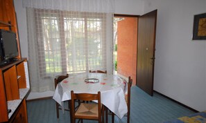 Ein kleiner Essbereich mit einem runden Tisch, der mit einer weißen Tischdecke bedeckt ist, Holzstühle, einem Fernseher auf einer Holzeinrichtung und Blick durch ein Fenster mit transparenten Vorhängen.