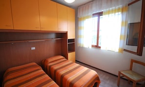 Ein Zimmer mit zwei Betten, einem Holzstuhl und einem Fenster mit Vorhängen.