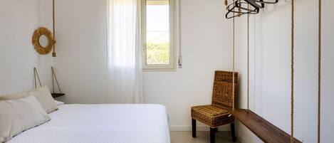 A bedroom with a bed, a chair, a window, and a wall-mounted rack.