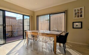 A dining area with a wooden table and chairs, large windows with curtains, and a view of a brick building outside. - Modern 3 Bed, 2.5 Bathroom House by the Beach (Seaford)