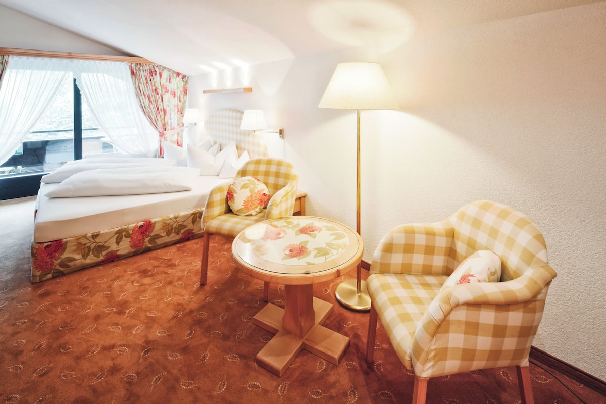 A hotel room with a bed, two checkered armchairs, a small round table, a floor lamp, and a window with curtains.
