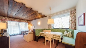 Une pièce chaleureuse avec un plafond en bois, un canapé vert, une table à manger recouverte d’une nappe à carreaux et une fenêtre ornée de rideaux à motifs floraux.