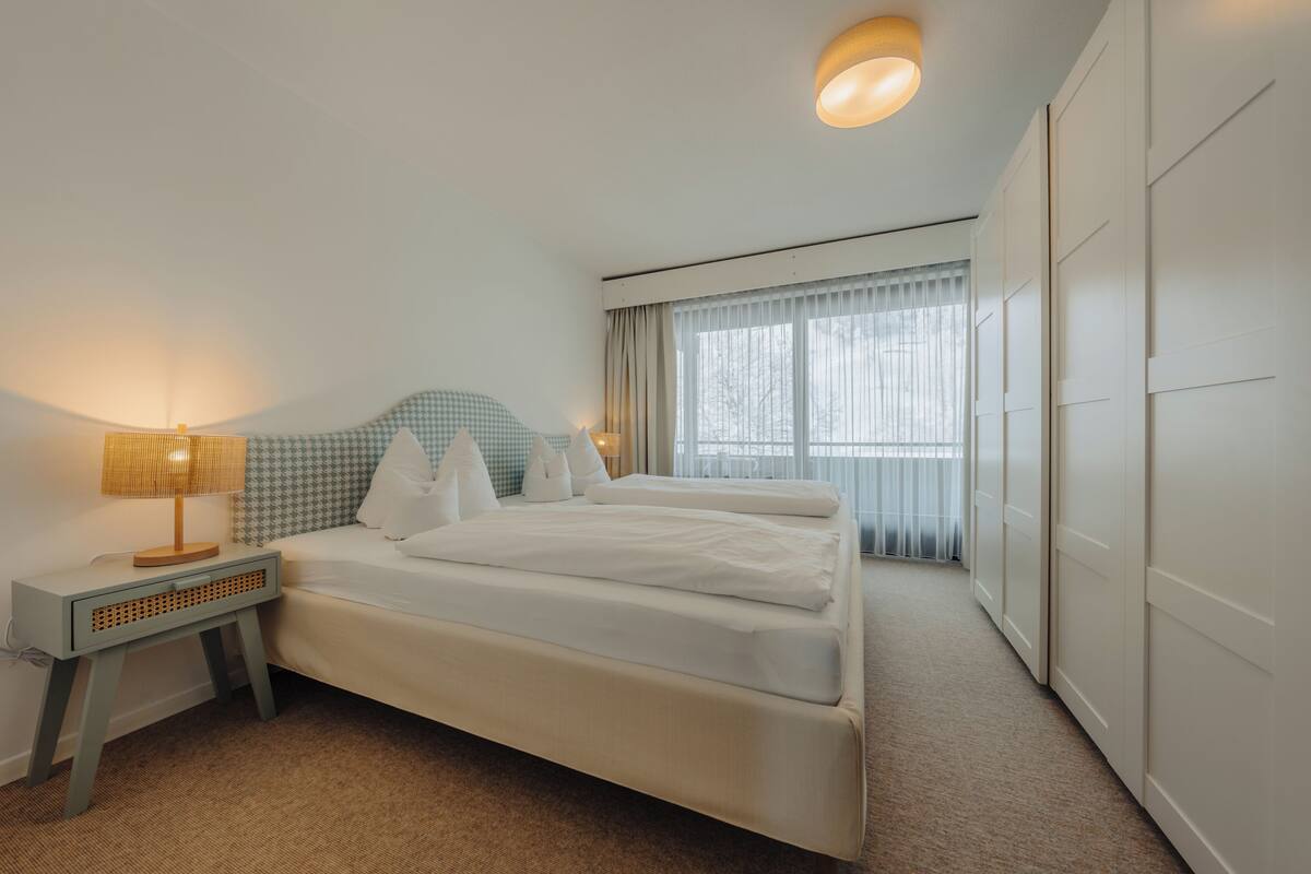 A hotel room with a bed, bedside table, lamp, and a view of the outdoors through the window.
