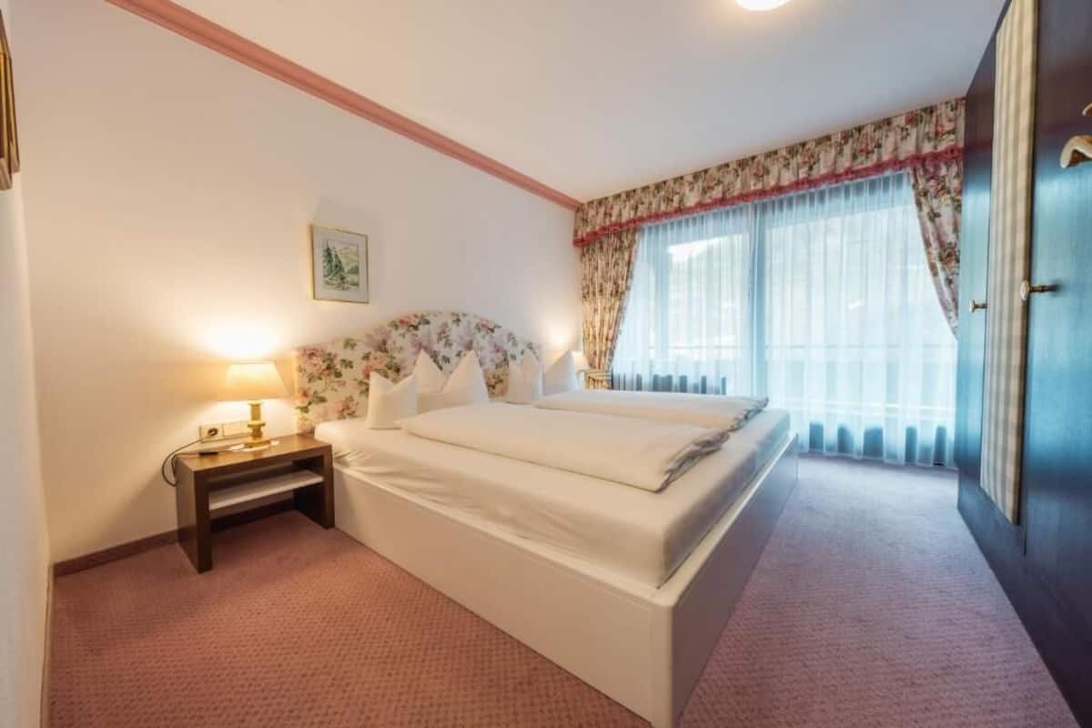 A hotel room with a bed, bedside table, lamp, floral headboard, and a window with curtains.