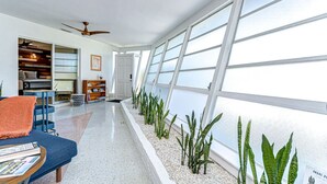 Un interno moderno con un lungo corridoio, una ventola a soffitto, una libreria e un'ampia finestra con piante.