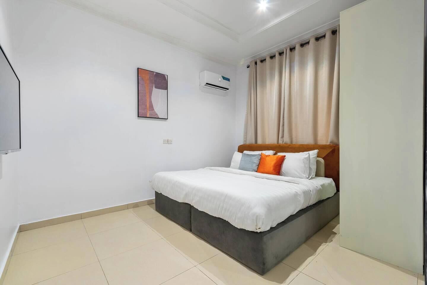 Un dormitorio con cama, aire acondicionado instalado en la pared, un cuadro enmarcado y una ventana con cortinas.