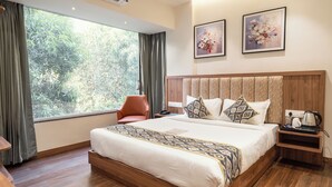 Habitación de hotel con un ventanal grande, una cama con ropa de cama estampada, cabecera de madera, mesita de noche con una tetera y dos cuadros enmarcados con motivos florales en la pared.