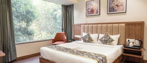 A hotel room with a large window, a bed with patterned bedding, a wooden headboard, a nightstand with a kettle, and two framed floral paintings on the wall.