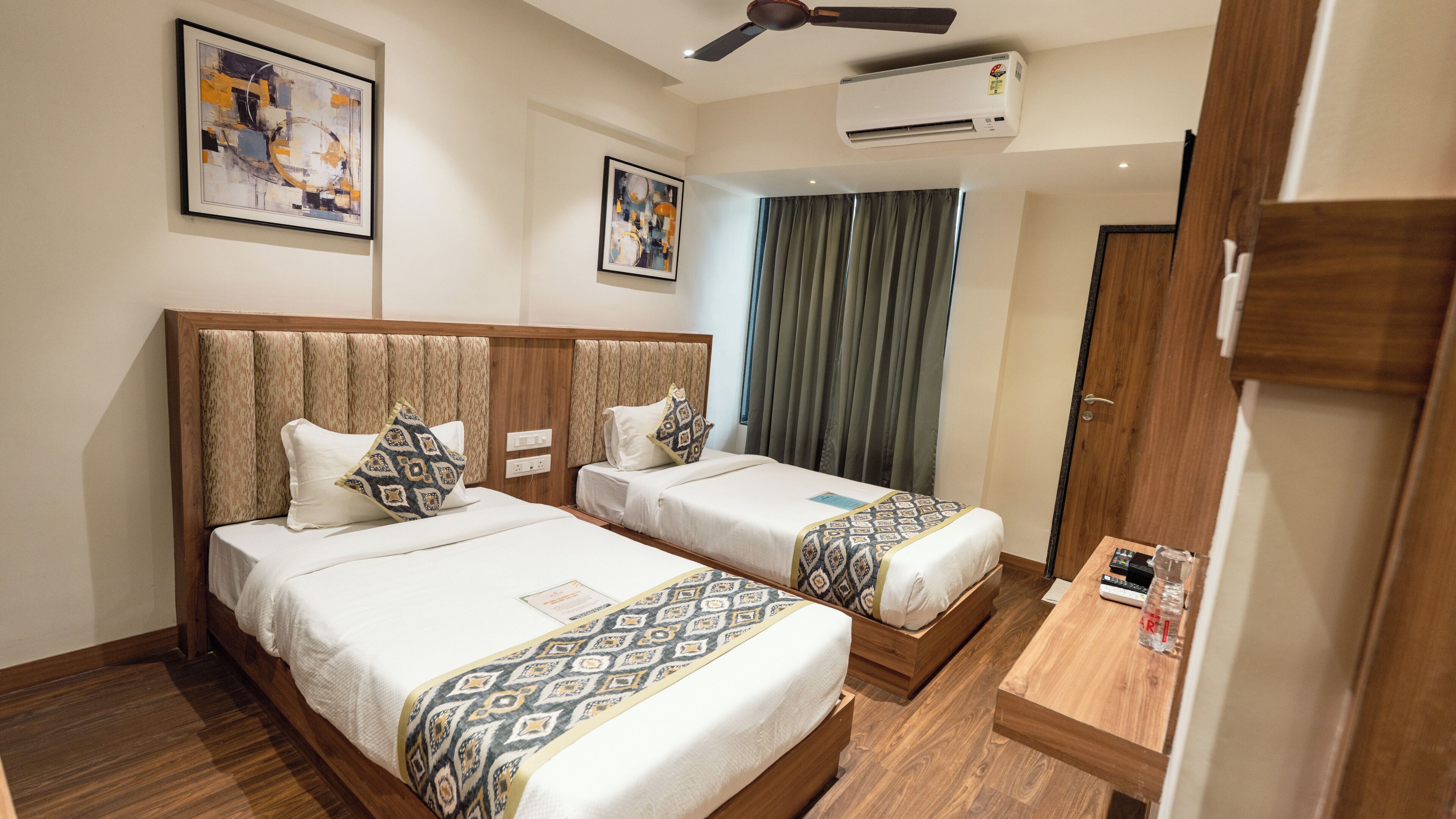 A hotel room with two beds, a ceiling fan, an air conditioner, and artwork on the walls.