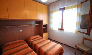 A room with two beds, a wooden chair, and a window with curtains. - Semi-detached House in Lignano Pineta (Lignano Sabbiadoro)
