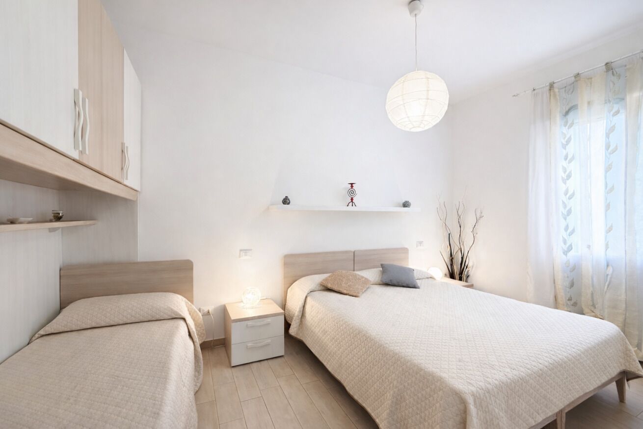 Une chambre moderne avec deux lits, une table de chevet avec une lampe, une étagère avec des objets décoratifs et un luminaire suspendu.