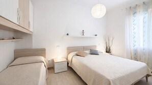 Un dormitorio moderno con dos camas, una mesita de noche con lámpara, una estantería con objetos decorativos y una lámpara colgante.