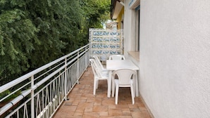 Pequeño patio al aire libre con una mesa y sillas blancas, barandilla blanca y vista a un árbol grande.